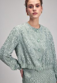 Ljus mintgrön broderad jacka med blommönster, rund halsringning, fem knappar och elastisk nederdel med något puffade ärmar.