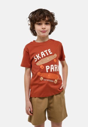 Garçon aux cheveux bouclés portant un t-shirt orange rouille "Skate Park" et un short kaki, debout avec une main dans la poche sur un fond blanc.
