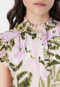 Blouse florale en rose clair avec des accents verts et violets, dotée d'un col volants, d'une fermeture à boutons et de manches courtes.