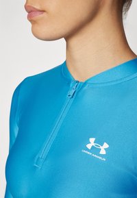 Plavi atletski top od glatke tkanine. Ima zatvaranje na patentni zatvarač na vratu i bijeli Under Armour logo na prednjoj strani.