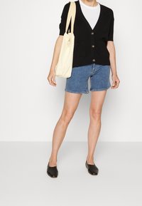 Svart kortärmad cardigan med knappar, vit t-shirt, jeansshorts och svarta slip-on skor. Bär en ljusbeige tote-väska.