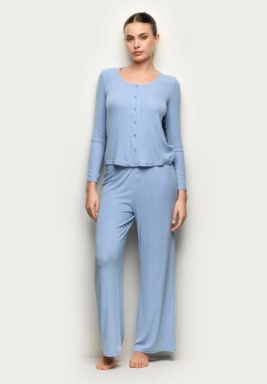 Kobieta stojąca na jednolitym tle, ubrana w jasnoniebieską, zapinaną na guziki bluzkę z długim rękawem oraz luźne spodnie w tym samym kolorze, boso.