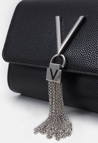 Primo piano di una borsa in pelle nera con texture, dotata di una chiusura a V in argento e una nappina a catena argentata che pende, su sfondo bianco.