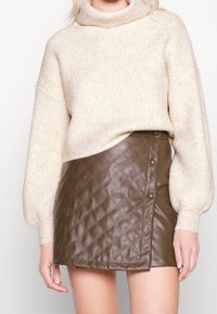 Kobieta ubrana w beżowy sweter z golfem o prążkowanej strukturze oraz brąskową, pikowaną skórzaną spódniczkę mini z detalami guziczków.