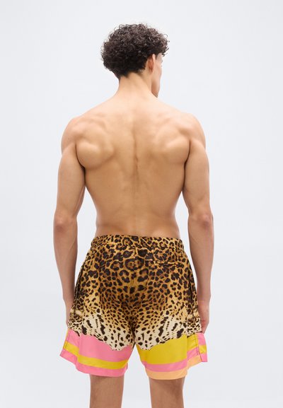 Άνδρας με σγουρά μαλλιά, γυμνή πλάτη προς τα εμπρός, φοράει πολύχρωμο μαγιό με τύπωμα λεοπάρδαλης και ροζ και κίτρινες ρίγες.