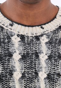 Gros plan d'un pull en tricot noir et blanc avec un motif torsadé, porté par une personne à la peau de ton moyen-foncé.