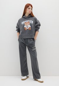 Felpa grigia con la scritta "Brooklyn" e grafica di un orsetto, abbinata a pantaloni della tuta larghi coordinati con un accento logo, indossati con sneaker bianche.