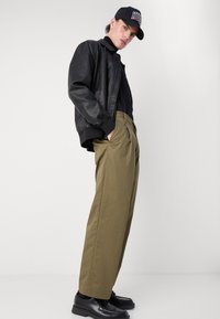 Veste bomber en cuir noir, pull noir ajusté, pantalon large vert olive, chaussures noires et casquette noire avec lettrage blanc.
