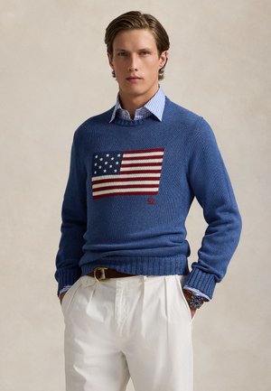 Mladý muž oblečený v modrom svetri s pleteným vzorom americkej vlajky, doplnený bielymi nohavicami a pruhovanou košeľou s golierom.