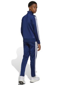 Tuta blu navy con strisce bianche, caratterizzata da un colletto alto e una giacca con cerniera. I pantaloni hanno polsini elasticizzati. Indossata con scarpe da ginnastica bianche.