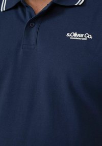Dunkelblaues Poloshirt aus strukturiertem Stoff mit einer Dreiknopf-Leiste und einem weißen gestreiften Kragen. Logo auf der linken Brust.