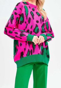 Pull rose oversize présentant un imprimé léopard noir et vert, avec un ourlet et des poignets côtelés verts, porté avec un pantalon vert.