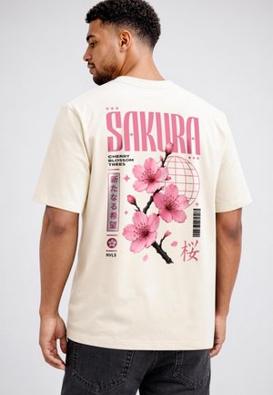 Man wearing cream T-shirt with large pink cherry blossom design and text "SAKURA" on the back, standing against plain background.