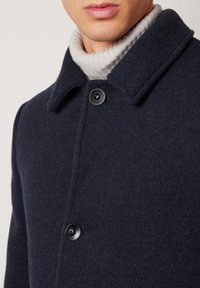 Marinblå ullkappa med texturerad yta, med krage, två stora svarta knappar och en lager av grå stickad polotröja under.