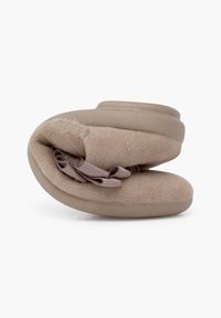 Zapatilla beige con parte superior de tela suave, forma redondeada, acentos elásticos y suela acolchada. Superficie texturizada con un ligero brillo.