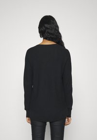 Pull en maille noire à manches longues et col rond, présentant un motif texturé et un ourlet légèrement incurvé à l'arrière.