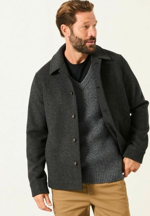Dunkelgraue Fischgrätjacke mit Kragen, Knopfverschluss, frontalen Taschen, getragen über einem hellgrauen gerippten Pullover und hellbraunen Hosen.