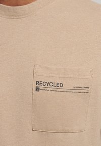 Camiseta de algodón beige con cuello redondo, que presenta un bolsillo en el lado izquierdo del pecho. El bolsillo tiene un texto impreso en negro que dice "RECICLADO" y información adicional.