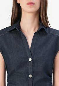 Mørk denimskjorte med krage, kort erme og sølvfargede knapper. Har synlig søm og en jevn tekstur.