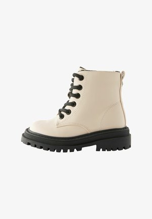 Beež nahast pahkluu saapad mustade nööpidega, ümmargune nina ja paks musta tallaga. Tagasisuunaline lukklukustus, millel on tekstureeritud viimistlus.