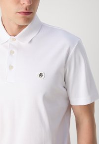Camisa polo branca feita de um tecido suave, com um pequeno logótipo bordado no lado esquerdo do peito, três botões no colarinho e mangas curtas.
