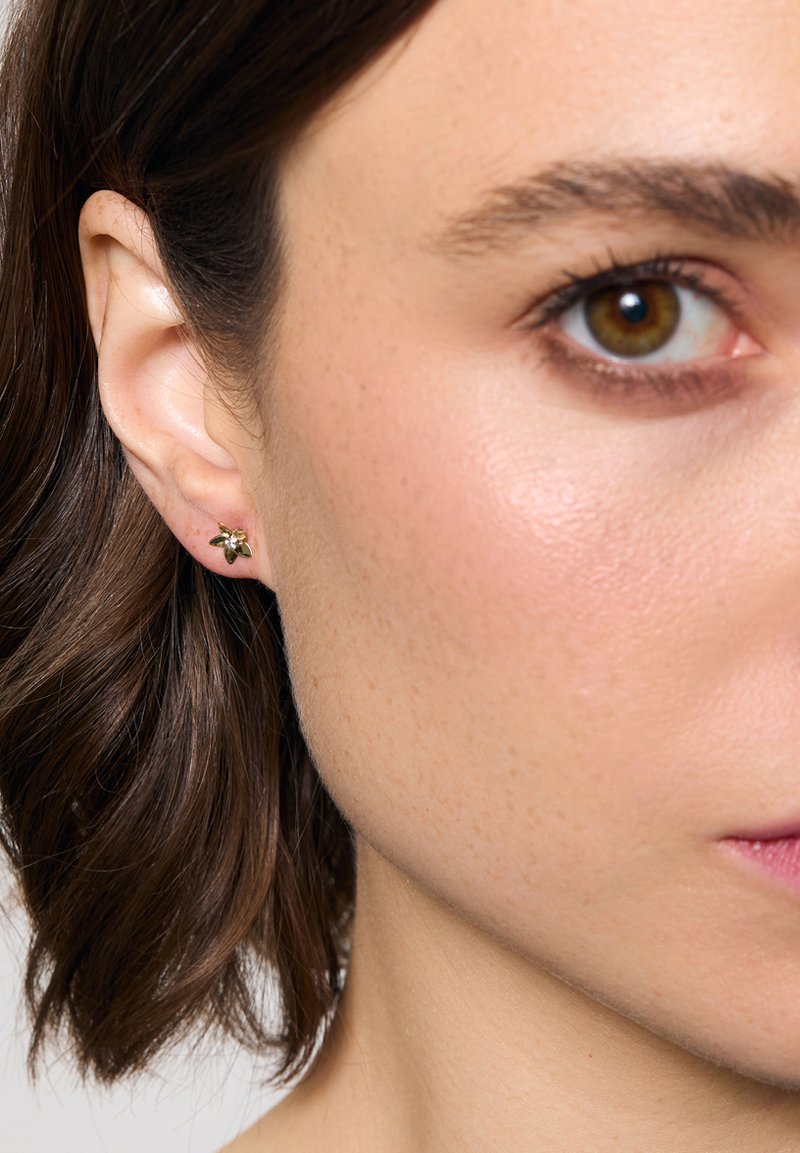 Złote metalowe kolczyki w kształcie kwiatu, ozdobione zielonymi liśćmi i małym różowym kamieniem w środku.