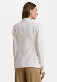 Blazer blanc ajusté avec un design cintré, des épaules structurées et des accents de boutons dorés sur les poignets, associé à un pantalon beige.