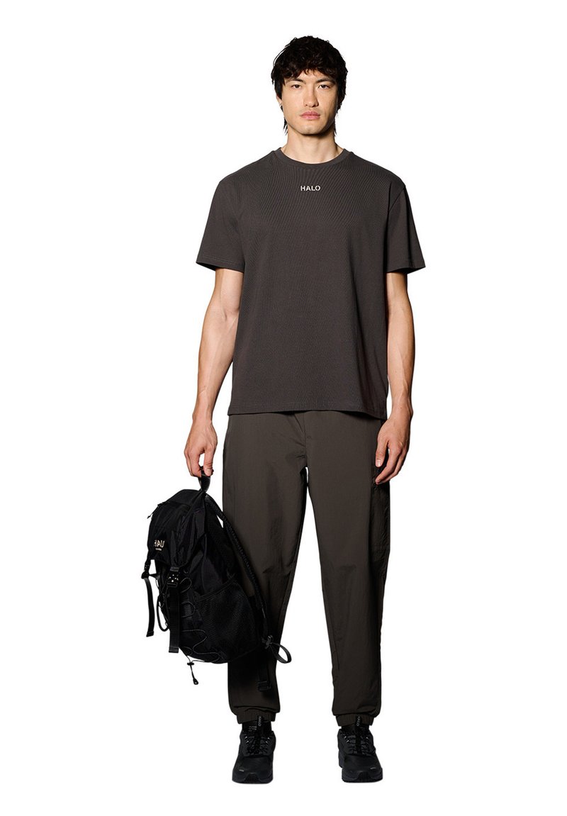 Svart texturerad t-shirt med "HALO"-logotyp, olivgröna cargo-byxor, svart mesh-ryggsäck och svarta sneakers, modell står rak.