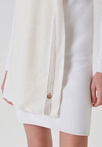 Sciarpa di misto lana color crema con texture a coste e accentuazione metallica lungo il bordo, drappeggiata su un vestito a maglia bianco con orlo aderente.