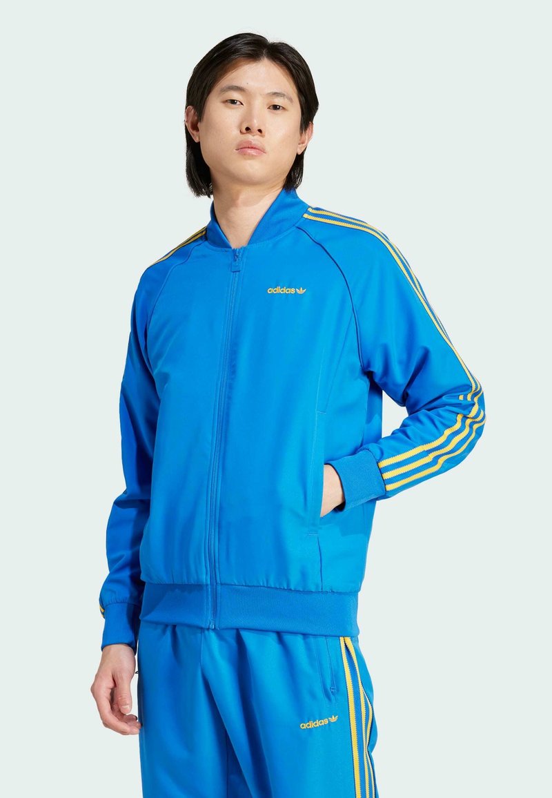 Plava jakna na zip od glatkog materijala, s žutim detaljima na ramenu i rukavima, s Adidas logotipom na prsima.