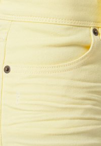 Keltainen farkkukangas, jossa on sileä rakenne, etutasku messingistä niitityksellä ja näkyvillä ompeluilla saumoissa.