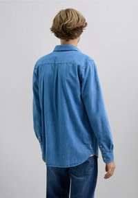 Camisa de mezclilla azul claro de manga larga, con cuello abotonado y dobladillo curvado. Presenta un yugo trasero con pliegues y puños abotonados.