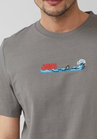 Szara bawełniana koszulka z czerwonym napisem "JAWS" oraz haftowanym wzorem płetwy rekina i niebieskiej fali. Okrągły dekolt i krótkie rękawy.