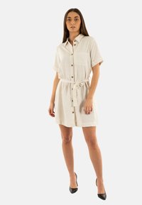 Abito camicia in lino beige con maniche corte, chiusura frontale con bottoni e vita con lacci. Presenta una tasca sul petto e una texture morbida. Indossato con tacchi neri.