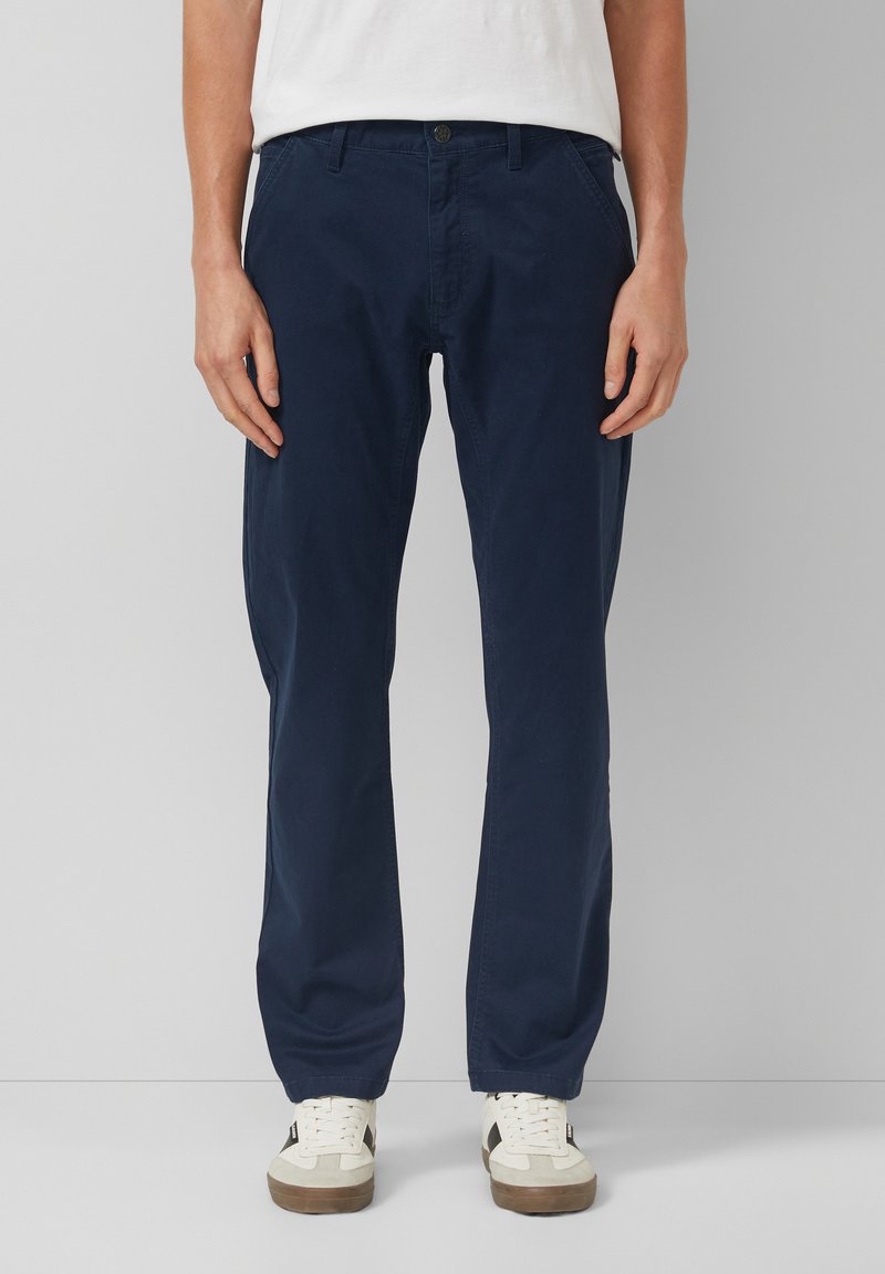 Uomo che indossa pantaloni blu navy a gamba dritta, sneakers bianche con dettagli neri e una camicia bianca, in piedi contro uno sfondo grigio.