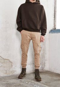 Sweat-shirt à capuche marron avec une poche avant, associé à un pantalon cargo beige clair avec des poches latérales et des chevilles ajustées, complété par des bottes foncées.