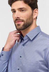 Camicia a scacchi blu e bianca con colletto alla francese, dettagli con bottoni e tessuto testurizzato, indossata da un uomo che si tocca il collo.