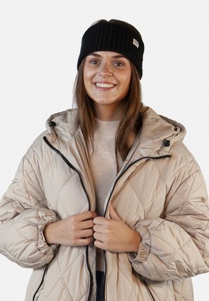 Giacca trapuntata beige con cappuccio, dotata di una zip nera e polsini elasticizzati, indossata su un maglione beige chiaro e un berretto di maglia nero.