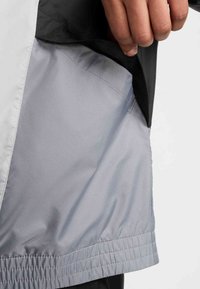 Αδιάβροχο μπουφάν με μαύρο εξωτερικό στρώμα και εσωτερική επένδυση ανοιχτού γκρι. Λείο ύφασμα με ελαστικό τελείωμα και ραφές λεπτομέρειας.