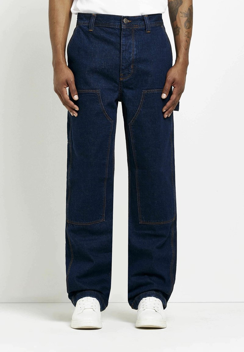 Mörkblå denimbyxor med avslappnad passform, med stora framfickor och synliga sömdetaljer. Bärs med vita sneakers.