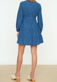 Donna che indossa un vestito blu a strati con maniche lunghe e zoccoli beige con tacco, voltata di spalle su un pavimento beige con uno sfondo bianco semplice.