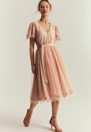 Kobieta ubrana w różową, sięgającą do kolan sukienkę z tiulu z bufkami, haftowanym kwiatowym wykończeniem, zapiętym gorsetem i przewiązanym paskiem w talii, w połączeniu z obcasami.