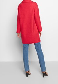 Mujer vestida con un abrigo rojo brillante hasta la rodilla, jeans azules y sandalias de tacón negro, de pie contra un fondo blanco.