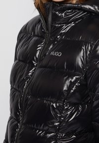 Czarna pikowana kurtka z błyszczącym wykończeniem, zamkiem błyskawicznym, wysokim kołnierzem i przeszywanymi panelami. Na lewej stronie znajduje się logo „HUGO”.