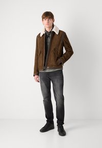 Chaqueta de ante marrón con un cuello de borrego blanco, dos bolsillos frontales, combinada con una camisa gris, jeans grises y zapatillas negras.