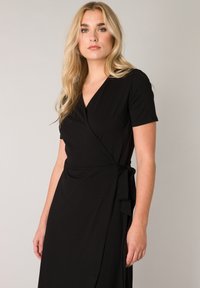 Zwarte wikkeljurk met korte mouwen, V-hals en strikdetail in de taille. Gemaakt van gladde stof met een расслаблен fit en draping.