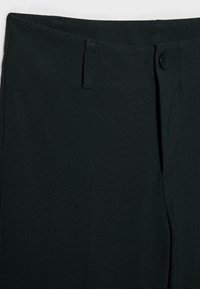 Gros plan sur la ceinture d’un pantalon noir avec passants pour ceinture, fermeture par bouton et texture du tissu visible.