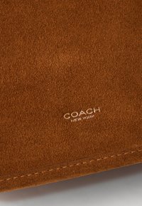 Brązowy zamsz z miękką fakturą, z wytłoczonym logo "COACH NEW YORK" w kolorze srebrnym w prawym dolnym rogu.