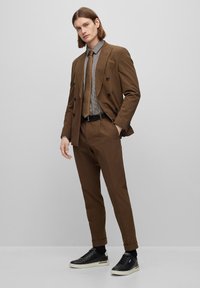 Giacca doppiopetto marrone sopra una camicia a quadri e cravatta marrone. Pantaloni slim-fit e sneaker nere con suola bianca.