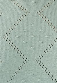 Lichtgroene gebreide stof met diamantpatronen, kleine gestructureerde stippen en contrasterende bruine accenten in de stiksels. Zachte textuur.